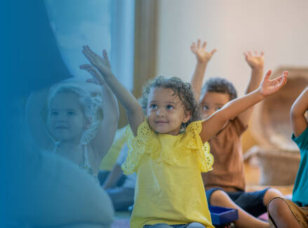 early childhood students in a classroom raising their hands.
